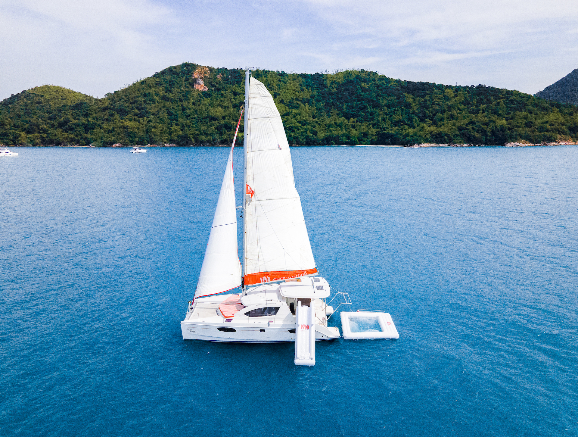 Rose sailing catamaran with its sail raised, seen from above near an island